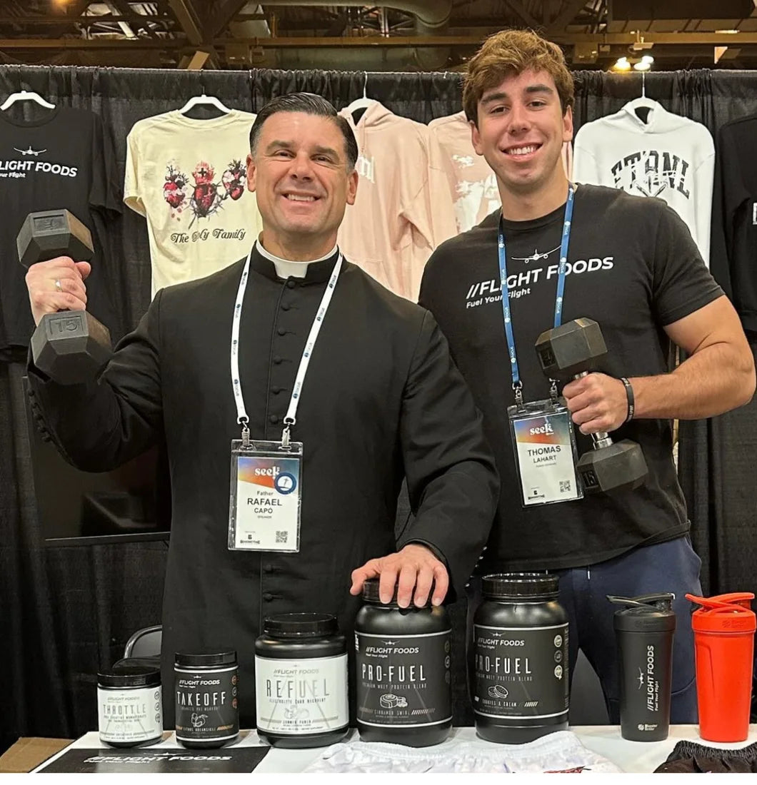 Fr. Rafael Capó curling a dumbbell beside founder Thomas Lahart at the Flight Foods booth at SEEK